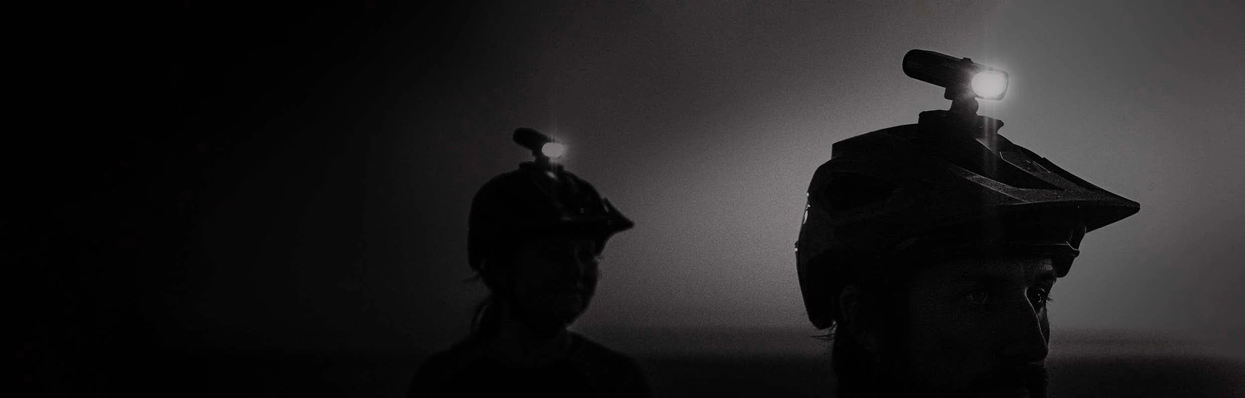 Helmet-mounted bike lights on two helmets worn by people in a dark setting, highlighting the powerful illumination and secure mounting.