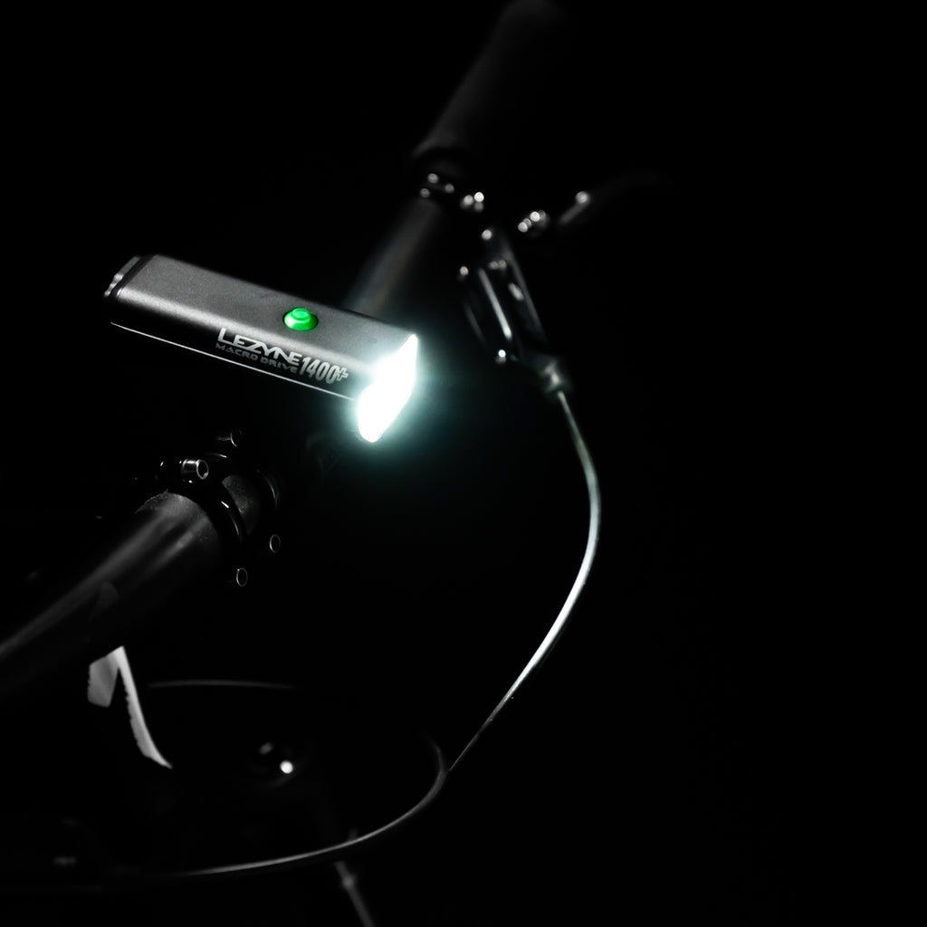 Top view of MACRO DRIVE 1400+ front LED bike light, mounted on gravel bike handlebar, showcasing a powerful light beam and green power indicator.
