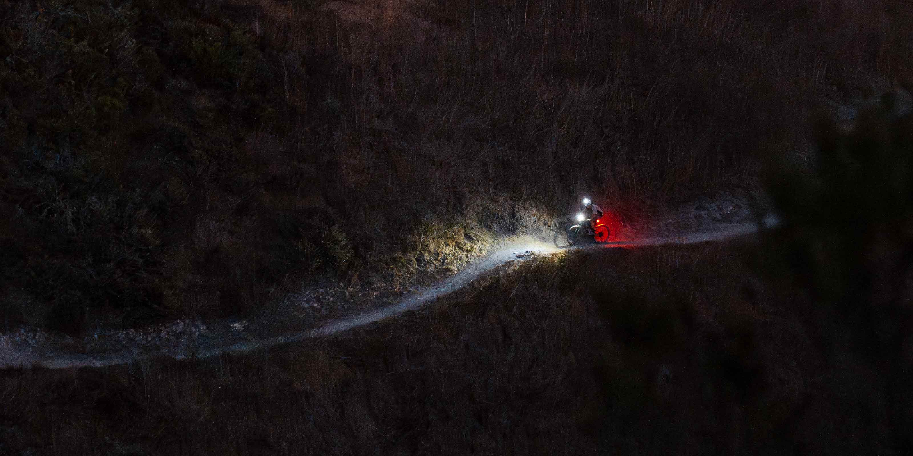 Mountain biker in the dark on the side of a hill with bright lights illuminating the trail ahead