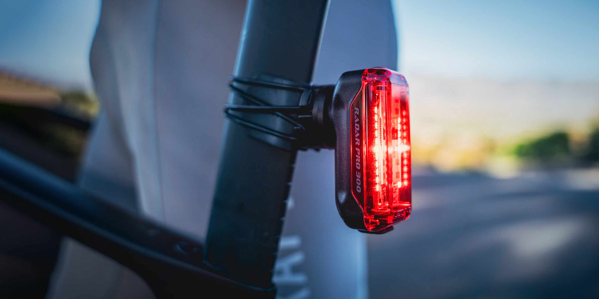 A close-up shot of a Radar Pro 300 rear light mounted on a dark frame, with the red LED light illuminated against a blurred outdoor background.